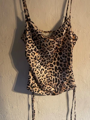 Leopardmönstrad topp med tunna band - Säljer en snygg leopardmönstrad topp med tunna axelband och draperad urringning. Toppen har justerbara knytband i sidorna och är gjord i ett glansigt, slätt material som känns lyxigt. Perfekt för dig som vill sticka ut med ett djurmönster. Ingen aning om vart det är ifrån men skulle tro på H&M