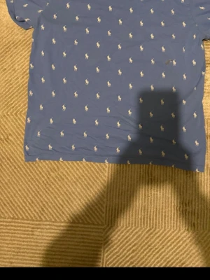 Blå t-shirt från Polo Ralph Lauren - Snygg blå t-shirt från Polo Ralph Lauren med små vita loggor tryckta över hela tröjan. Klassisk passform med rund hals och korta ärmar. Perfekt för dig som gillar stilrena plagg med en twist. Materialet känns mjukt och bekvämt mot huden.