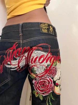Christian Audigier jeans med brodyr - Säljer ett par mörkblå jeans från Christian Audigier med stor broderad text, vita vingar och röda rosor på bakfickan. Jeansen har detaljer i rött, grönt och vitt samt en unik, edgy design som verkligen sticker ut.