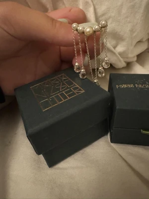 Maria Nilsdotter örhänge med pärlor - Unikt örhänge från Maria Nilsdotter i silverfärg med fem vita pärlor upptill och sex hängande kedjor med droppformade klara stenar. Snygg och modern design som sticker ut och ger en lyxig känsla.