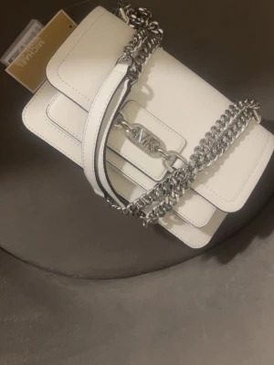 Vit handväska från Michael Kors - Snygg vit handväska från Michael Kors i slätt läder med silverfärgade kedjedetaljer och MK-logga framtill. Väskan har både axelrem och kedjerem, samt stilrena sömmar och en rektangulär form. Perfekt accessoar för att lyfta din outfit. Ny pris 3300kr