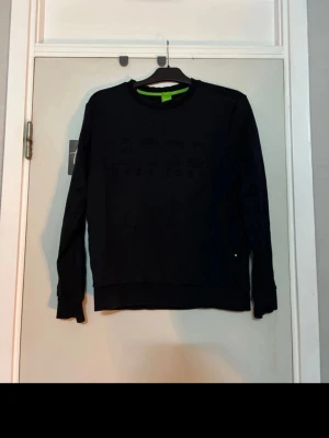 Svart sweatshirt från Hugo Boss - Snygg svart sweatshirt från Hugo Boss med diskret broderad logga på bröstet. Tröjan har rund halsringning, långa ärmar och ribbade muddar. Perfekt för en clean och stilren look. Materialet känns mjukt och bekvämt.