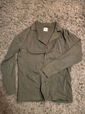 Olivgrön cardigan från John Henric - Stilren olivgrön cardiganjacka från John Henric med klassisk krage, två stora framfickor och knappstängning. Tillverkad i mjukt, tunt stickat material som ger en avslappnad men ändå snygg look. Perfekt att slänga över en t-shirt eller skjorta.