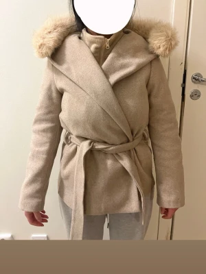 Beige kappa med fuskpäls och luva - Stilren beige kappa med knytskärp i midjan och stor luva kantad med fluffig fuskpäls. Kappan har omlottstängning och är tillverkad i ett mjukt, ull-liknande material. Perfekt för kyliga dagar och ger en trendig look. Möts hällre upp, vi finns i stockholm/södertälje.