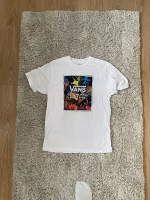 Vit Vans t-shirt med färgglatt tryck - Säljer en vit t-shirt från Vans med ett stort, färgglatt tryck på bröstet. Trycket har blommor och texten 'VANS OFF THE WALL SINCE 1966'. Klassisk passform och rund hals. Perfekt för dig som gillar streetwear och vill sticka ut lite extra.
