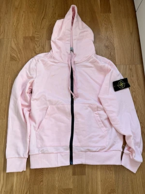 Ljusrosa hoodie från Stone Island - Ljusrosa hoodie från Stone Island med dragkedja framtill och klassisk svart logotyp-patch på vänster ärm. Hoodien har två fickor fram, ribbade muddar och en stor huva med dragsko. Tillverkad i mjukt bomullsmaterial som känns skönt mot huden. den är strl S och är helt ny aldrig använd