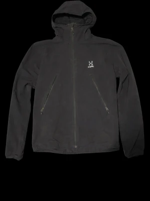 Svart vindjacka från Haglöfs - Svart vindjacka från Haglöfs med huva och två snedställda dragkedjefickor framtill. Jackan har minimalistisk design, diskret logga på bröstet och dragkedja hela vägen upp. Perfekt för blåsiga dagar och enkel att matcha med olika outfits.