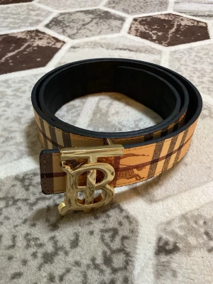 Beige och brun rutig Burberry-bälte - Snyggt bälte från Burberry i klassiskt beige och brunt rutigt mönster. Bältet har ett stort guldfärgat spänne med Burberrys ikoniska TB-logga. Perfekt accessoar för att lyfta din outfit med en lyxig touch.