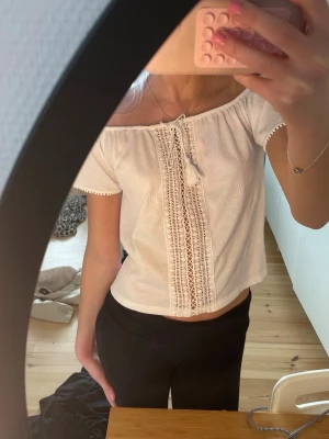 Vit offshoulder blus med spetsdetaljer - Superfin vit offshoulder blus med spets och knyt framtill. Blusen har korta ärmar med små bollar i kanten och är gjord i ett lätt bomullstyg. Perfekt för varma dagar och ger en romantisk vibe.