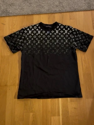 Svart  t-shirt med monogram - Säljer en svart t-shirt med klassiskt monogrammönster i vitt över axlar och bröst. T-shirten har rund hals, korta ärmar och är tillverkad i mjuk bomull. Perfekt för dig som vill ha en statement piece i garderoben.