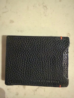 Svart läderplånbok från Tiger of Sweden - Snygg svart plånbok i äkta läder från Tiger of Sweden. Plånboken har en stilren, klassisk design med strukturmönstrad yta och små röda detaljer i hörnen. Perfekt storlek för kort och sedlar, och enkel att ha i fickan.