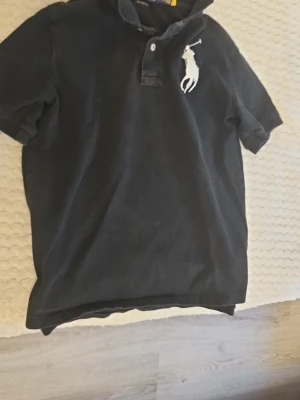 Svart pikétröja från Polo Ralph Lauren - Klassisk svart pikétröja från Polo Ralph Lauren med stor vit logga på bröstet. Tröjan har korta ärmar, krage och knappar framtill. Perfekt till jeans eller shorts för en clean och stilren grish style