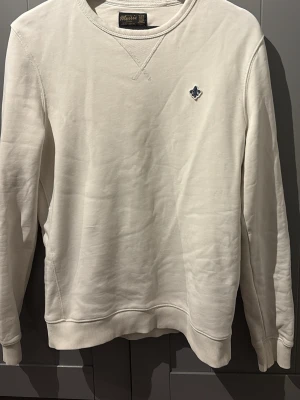 Morris tröja - Klassisk vit sweatshirt från Morris med rund halsringning och diskret blå logga broderad på bröstet. Tröjan har ribbade muddar vid ärmslut och nederkant, samt en mjuk bomullskänsla som gör den riktigt skön att bära.