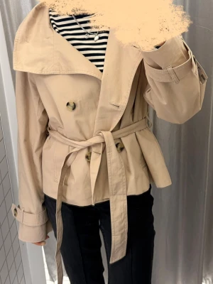 Kort beige trenchcoat från Bershka - Stilren beige trenchcoat från Bershka med dubbelknäppning, bred krage och knytskärp i midjan. Jag vill gärna byta till storlek xs eller s!