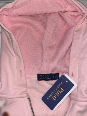 Ljusrosa hoodie från Polo Ralph Lauren - Snygg ljusrosa hoodie från Polo Ralph Lauren med dragkedja framtill och klassisk huva. Tröjan har två fickor på magen och den ikoniska blå loggan broderad på bröstet. Tillverkad i mjukt bomullsmaterial för en skön känsla. Perfekt för en avslappnad och trendig look.