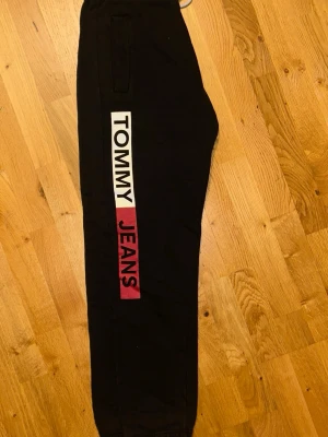 Svarta mjukisbyxor Tommy Jeans - Snygga svarta mjukisbyxor från Tommy Jeans med stort vitt och rött tryck längs ena benet. Byxorna har resår i benslut och midja samt fickor på sidorna. Perfekta för en avslappnad och trendig look.