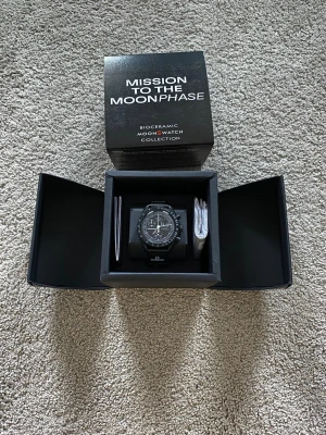 Omega x Swatch MoonSwatch svart klocka - Säljer en Omega x Swatch MoonSwatch Mission to the Moonphase i svart biokeramik. Klockan har en klassisk rund form, svart urtavla med vita detaljer och tachymeter-skala. Kommer i originalbox med manualer. Perfekt för dig som gillar ikoniska klockor.
