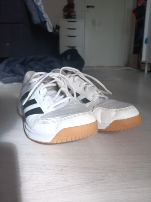 Vita Adidas sneakers med gummisula - Säljer ett par inneskor. vita Adidas sneakers med svarta ränder på sidorna och ljusbrun gummisula. Skorna har meshdetaljer på ovandelen och vita skosnören. 