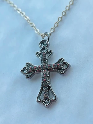 By prisförslag- Silverfärgat halsband 45 cm med korshänge - Snyggt halsband i silverfärg med ett dekorativt korshänge. Korset har små glittrande stenar infattade längs hela korset och fina detaljerade kanter. Kedjan är tunn och elegant, perfekt för att ge en stilren touch till din outfit.