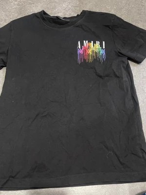 Svart AMIRI t-shirt med regnbågstryck - Snygg svart t-shirt från AMIRI med regnbågsfärgat tryck och droppande effekt på bröstet. Klassisk rund halsringning och korta ärmar. Tillverkad i mjuk bomull för en skön känsla och stilren look.