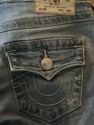 True Religion ljusblå joey low rise flare jeans - Säljer ett par ljusblå joey low rise flare jeans från True Religion med ikoniska broderade bakfickor och dekorativa knappar. Jeansen har normal midja och klassisk femficksdesign. Materialet är mjukt denim i bomull med lätt tvättad look. Jeansen användes ett par gånger och är i bra skick 