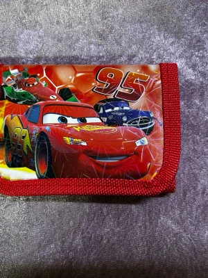 Röd Cars plånbok med motiv - Cool röd plånbok med motiv från Disney Pixar Cars och Lightning McQueen. Plånboken har färgstarka tryck, röda kanter och är tillverkad i slitstark textil och plast. Perfekt för dig som gillar bilar och vill ha en unik accessoar i fickan.