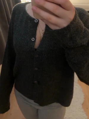 Mörkgrå mysig kofta med knappar - Mysig mörkgrå kofta med rund halsringning och knäppning framtill. Koftan har en lite luddig, stickad känsla och långa ärmar. Perfekt att slänga på sig över en topp för en avslappnad look.