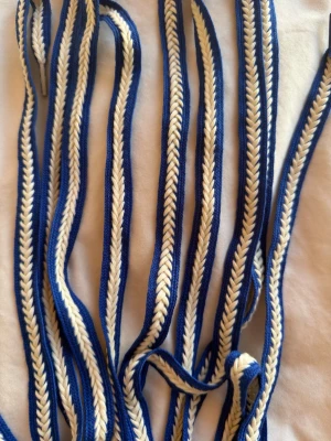 Blåvitt flätat tygskärp - Säljer ett blåvitt flätat tygskärp med fiskbensmönster. Skärpet är smalt och flexibelt, perfekt för att ge en cool touch till din outfit. Passar till jeans, kjol eller klänning och är lätt att knyta i olika stilar. Skosnören!