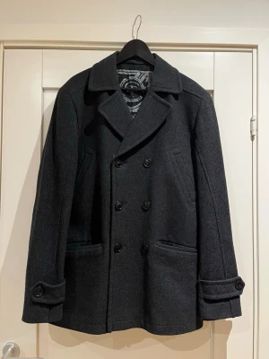 Jacka - Stilren mörkgrå/svart skepparkavaj med dubbelknäppning och bred krage. Kavajen har två snedställda fickor fram och dekorativa knappar vid ärmsluten. Från märket Cottonfield Tillverkad i en tjock ullmix som håller värmen. Fodret har ett mönster som ger extra karaktär. Tveka inte att fråga!! Kom med bud!!!