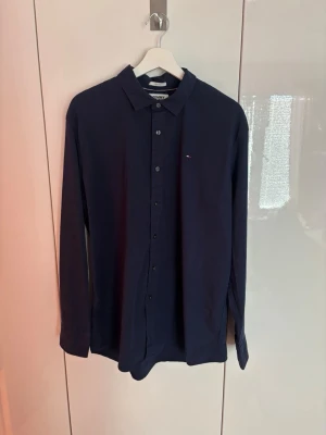 Mörkblå skjorta från Tommy Hilfiger - Klassisk mörkblå skjorta från Tommy Hilfiger med diskret logga på bröstet. Skjortan har lång ärm, knappar framtill och en stilren krage. Perfekt till både jeans och chinos för en clean look. Är XL men sitter som L