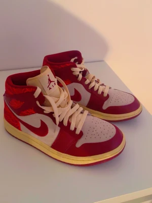 Air Jordan 1 Retro röd/vit - Snygga Air Jordan 1 Retro sneakers i rött och vitt med beige detaljer. Klassisk high-top siluett, perforerad tåbox och ikonisk Swoosh på sidan. Skorna har röda snören och gulaktig sula. Perfekta för dig som vill sticka ut med stil.