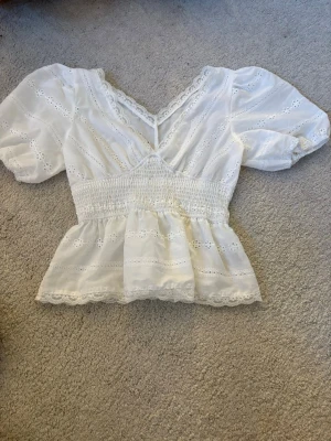 Vit blus med spets och v-ringning - Söt vit blus med v-ringning, puffärmar och smock i midjan. Blusen har fina spetsdetaljer och broderade blommor, samt volangkant nedtill. Passar väldigt fint med ett par kostymbyxor eller ett par jeans. Aldrig använd och vet tyvärr inte vart den är ifrån.