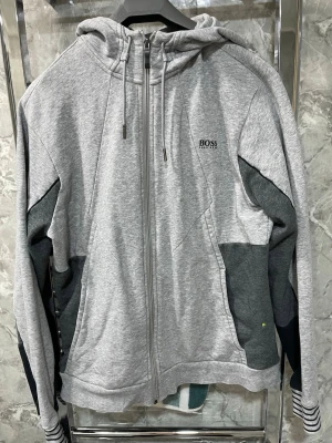 Grå hoodie från Hugo Boss - Snygg grå hoodie från Hugo Boss med dragkedja och huva. Tröjan har mörkgrå detaljer på ärmarna och sidan, samt BOSS-logga broderad på bröstet. Tillverkad i mjukt sweatshirtmaterial, perfekt för en avslappnad och trendig look.