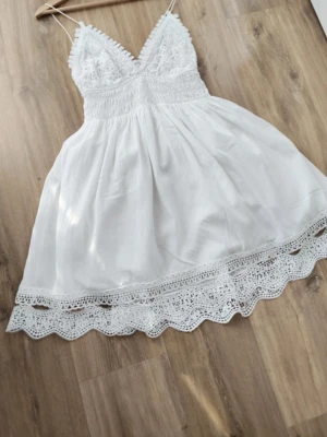  Ny Vit spetsklänning med v-ringning - Säljer en kort vit klänning med v-ringad spets upptill och tunna axelband. Klänningen har ett smockat liv och vacker spetsdetalj längs nederkanten. Perfekt för sommarens alla tillfällen, lätt och luftig i bomull.
