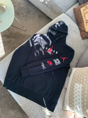 Svart Jordan x Nike hoodie - Snygg svart hoodie från Jordan med stor röd Jumpman-logga och texten 'JORDAN' på bröstet. Detaljer som Nike-logga, nummer 23 och signatur på ärmen. Klassisk känguruficka och huva. Perfekt för dig som gillar streetwear och sportig stil. Storlek XL, 13-15 år, 163 - 175 cm. 