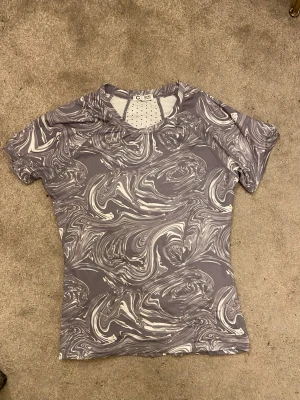 Grå sporttröja med marmor-mönster - Snygg sporttröja från Cubus i grått med vitt marmor-mönster. Tröjan har korta ärmar, rund hals och meshdetaljer upptill för extra ventilation. Tillverkad i ett stretchigt funktionsmaterial som passar perfekt till träning.