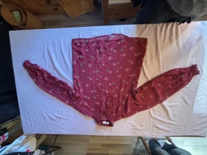 Röd blommig blus med knytband - Snygg röd blus med blommönster i röd och grönt. Blusen har långa ärmar med resår vid handlederna och ett dekorativt knytband vid halsen. Luftigt material som ger en avslappnad och trendig look.