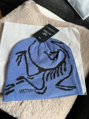 Blå mössa från Arc'teryx med tryck - Säljer en blå mössa från Arc'teryx med svart grafiskt tryck av deras ikoniska fossil-logga. Mössan är stickad i mjukt material och har en klassisk, något slouchy passform. Perfekt för dig som gillar stilrena accessoarer med en sportig touch.