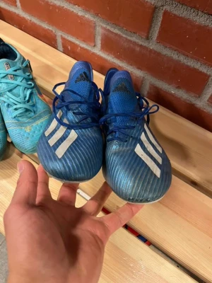  Adidas fotbollsskor  - Säljer ett par blå fotbollsskor från Adidas med klassiska vita ränder på sidan. Skorna har snörning och slitstark ovandel i syntetmaterial samt fasta dobbar för spel på gräs. Perfekta för dig som vill ha bra grepp och snygg stil på planen. Lite slitage 40,5 kan använda 