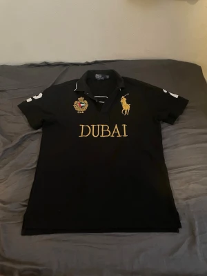 Svart polotopp från Ralph Lauren Dubai - Snygg svart polotopp från Ralph Lauren med guldfärgad DUBAI-text och logga framtill samt broderad patch. Stort nummer 2 på ryggen och vita detaljer på ärmarna. Klassisk krage och korta ärmar, perfekt för en sportig och trendig look.
