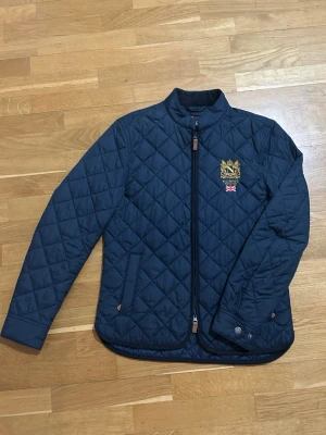 Mörkblå quiltad jacka från Morris - Säljer en stilren mörkblå quiltad jacka från Morris med broderad logga och brittisk flagga på bröstet. Jackan har dragkedja framtill och en klassisk krage. Perfekt för en elegant look.