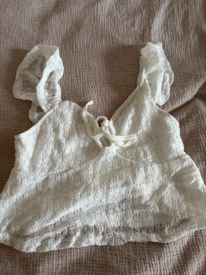 Vit spets topp med rosett - Söt vit topp i spets med kort, croppad passform och dekorativ rosett framtill. Toppen har breda axelband i spets och en lätt v-ringad urringning. Perfekt för dig som gillar romantisk och feminin stil.