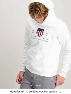 Vit hoodie från GANT med logga - Snygg vit hoodie från GANT med broderad logga och sköld på bröstet. Klassisk huva och stor magficka framtill. Tillverkad i mjukt material som känns skönt mot huden. Perfekt för en avslappnad och stilren look. Nypris 900kr