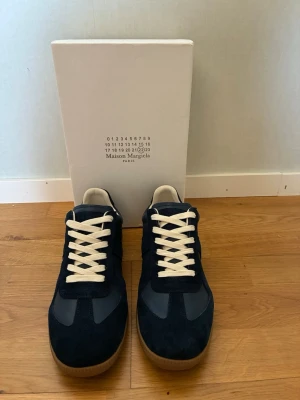 Maison Margiela sneakers - Snygga sneakers från Maison Margiela i mörkblå mocka och läder med vita skosnören och klassisk gummisula. Låg modell med stilren design och diskreta detaljer. Perfekta för dig som gillar minimalistisk och trendig stil.
