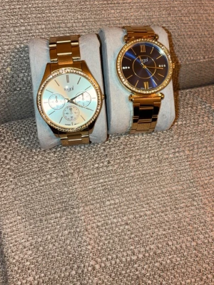 Två guldiga klockor från Regal - Säljer två stilrena klockor från Regal med guldigt metallarmband och glittrande stenar runt urtavlan. Den ena har svart urtavla med romerska siffror och den andra har ljus urtavla med flera små visare. Perfekta accessoarer för att lyfta din outfit.