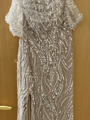 Långklänning med paljetter och slits - Elegant långklänning i beige med silverfärgade paljetter och pärlor i ett glittrigt mönster. Klänningen har korta ärmar, slits framtill och en vacker omlottliknande överdel. Perfekt för bal eller festliga tillfällen.