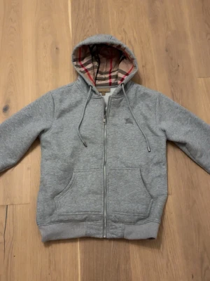 Grå zip hoodie från Burberry - Snygg grå hoodie med dragkedja från Burberry. Klassisk rutig huva, broderad Burberry-logga på bröstet och två stora fickor framtill. Mjuk bomullsblandning och justerbar huva med snören. Perfekt för en avslappnad och stilren look. den passar även M