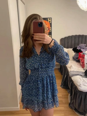 Blå mönstrad kortklänning med volang - Säljer en blå kortklänning med vitt mönster och volang nertill. Klänningen har långärmade transparenta ärmar, v-ringning och knytband vid halsen. Tillverkad i ett lätt och luftigt material, perfekt för varmare dagar.