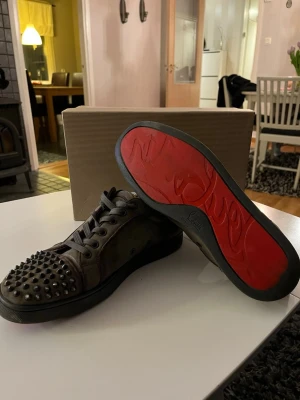 Christian Louboutin gråa spikes sneakers - Säljer ett par gråa sneakers från Christian Louboutin med ikoniska röda sulor och svarta nitar över tån. Skorna har lågt skaft, snörning och är tillverkade i mocka och läder. Perfekta för dig som vill sticka ut med en edgy och lyxig stil.