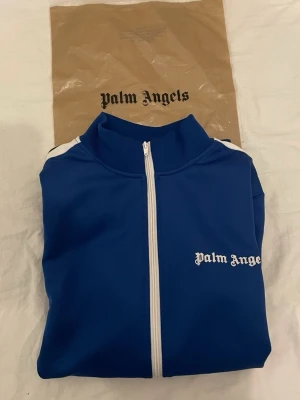 Blå track jacket från Palm Angels - Snygg blå track jacket från Palm Angels med vita detaljer och dragkedja framtill. Jackan har vita ränder längs ärmarna och logga på bröstet. Perfekt för en sportig och trendig look. Tillverkad i ett mjukt och stretchigt material.